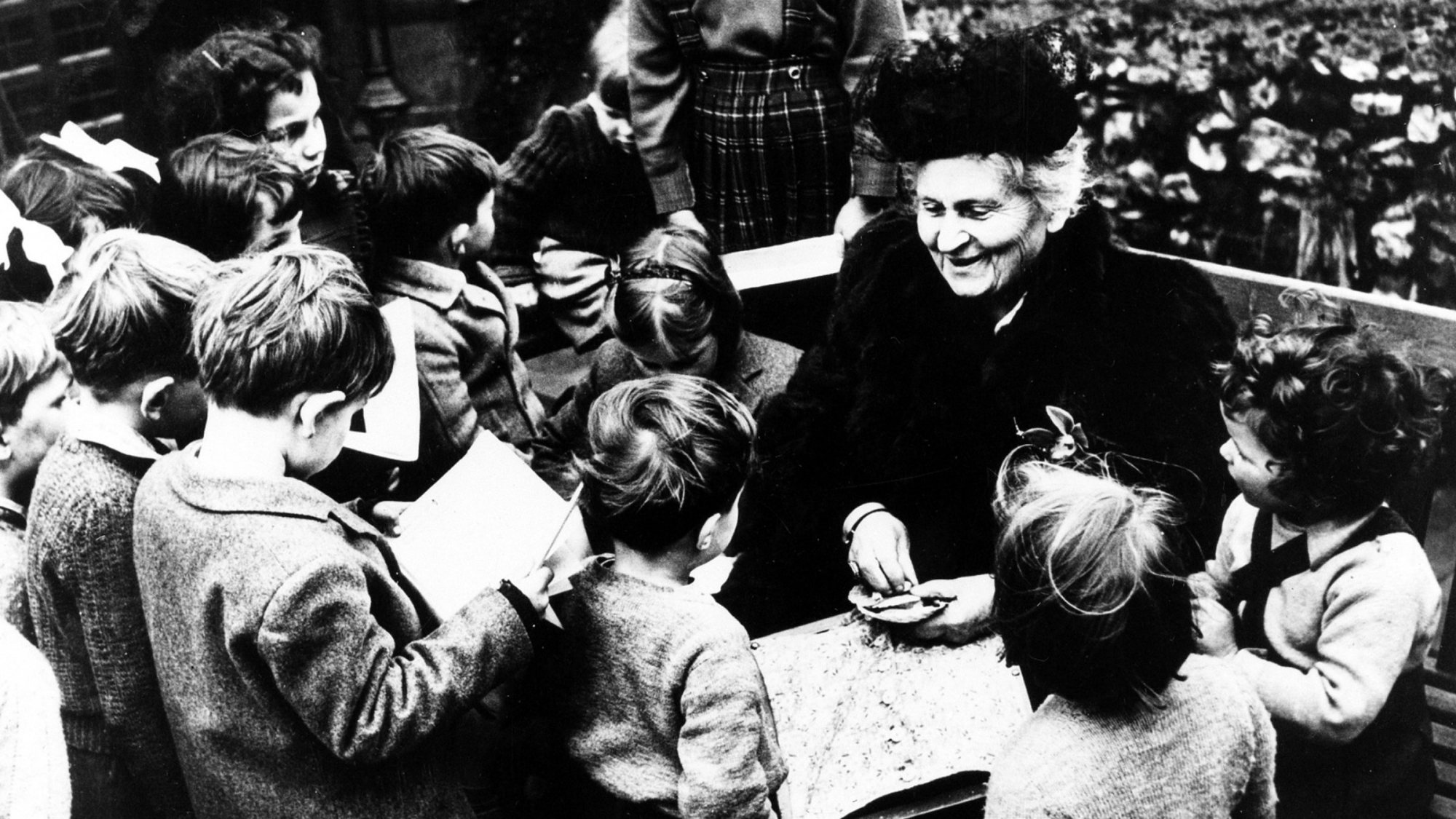 Historic photograph of Maria Montessori surrounded by young children.