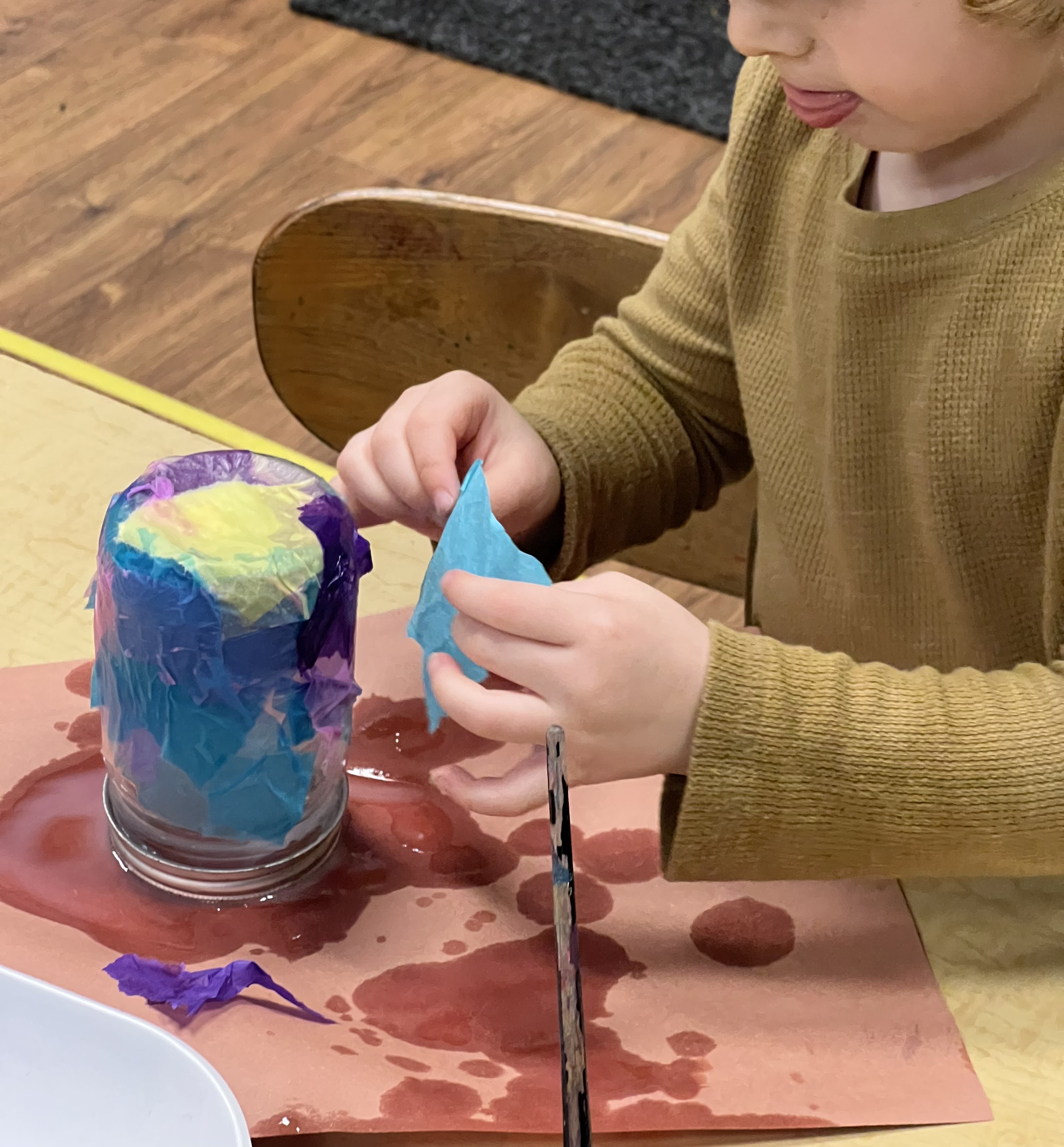 Preschool-aged child decorating a glass jar with tissue paper.
