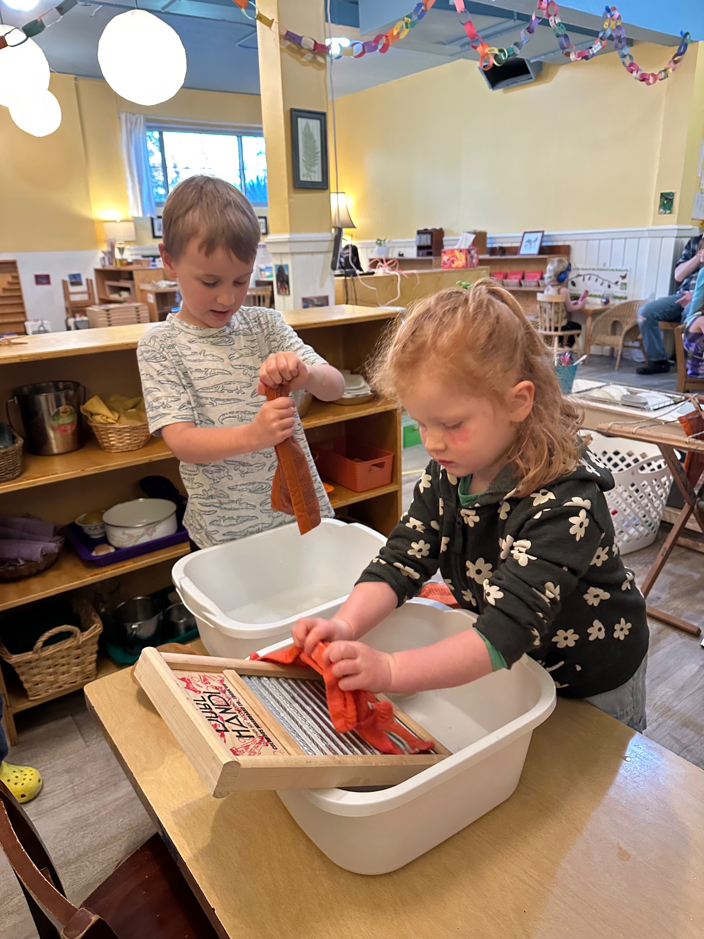 A preschool-aged boy and girl doing a cloth-washing work. The boy is squeezing water out of a cloth, the girl is washing a cloth on a washing board.