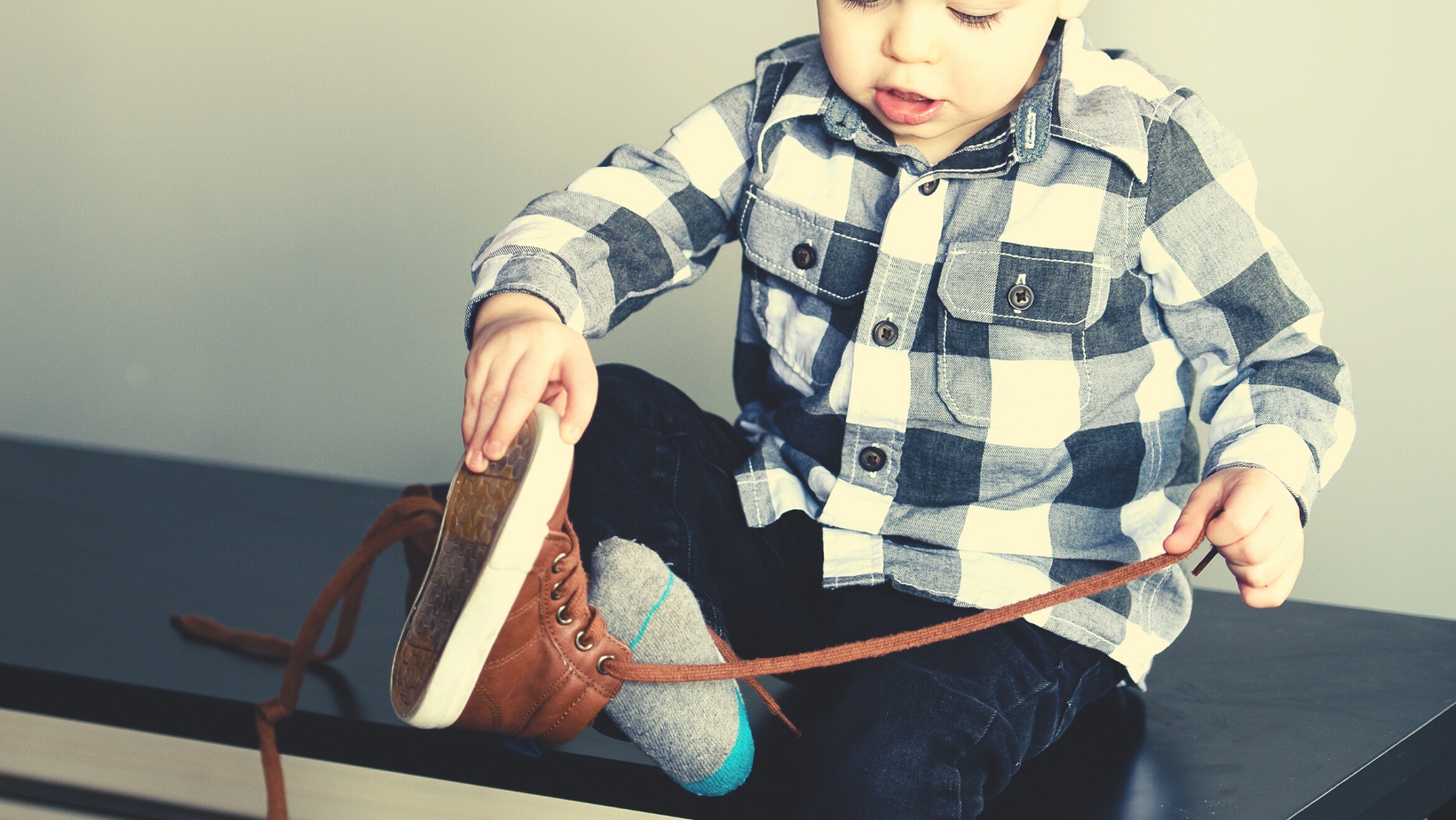 A preschool-aged boy sits on a bench trying to put his shoe on.