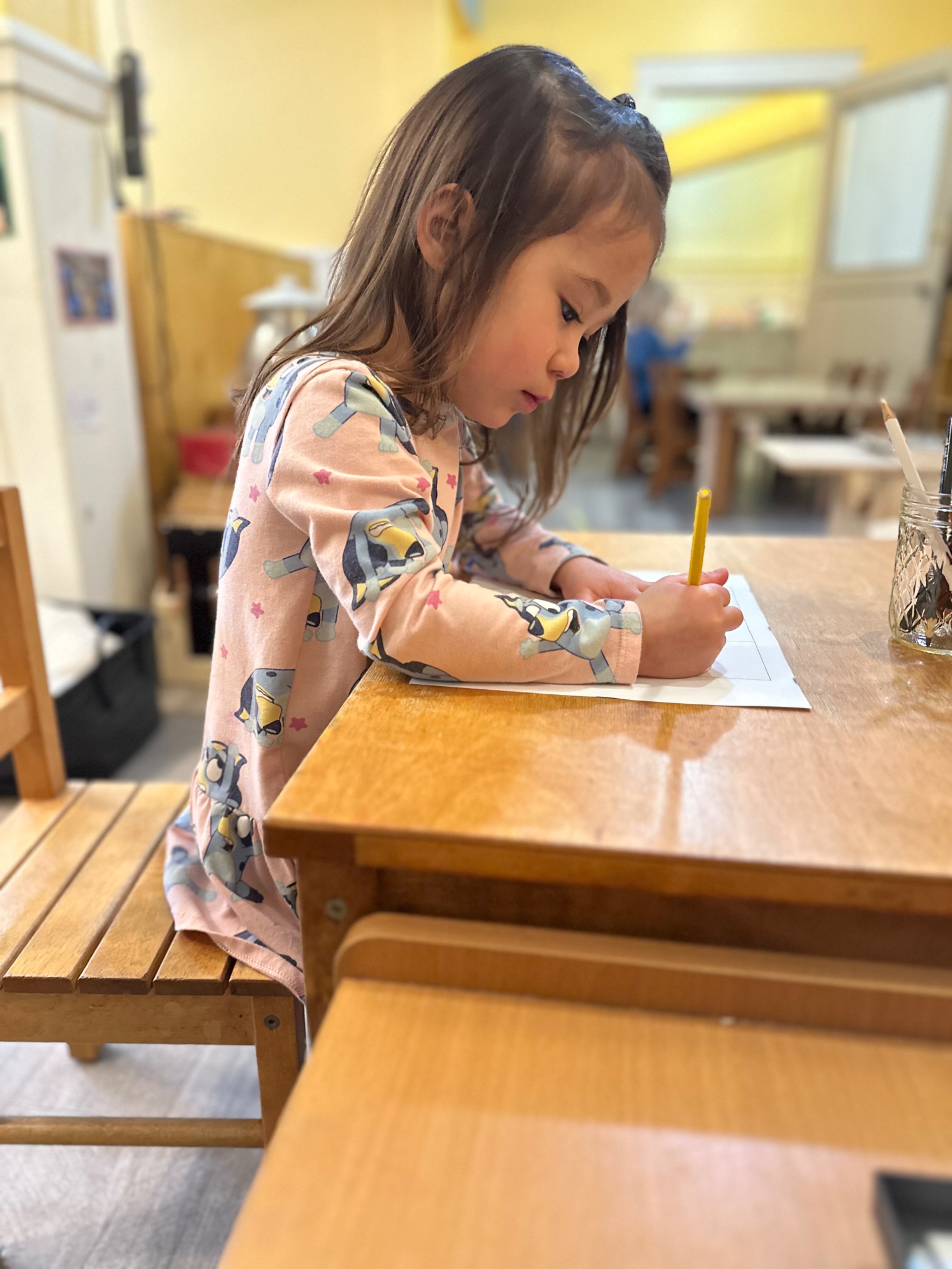 A preschool-aged girl at a table drawing on paper with colored pencils.