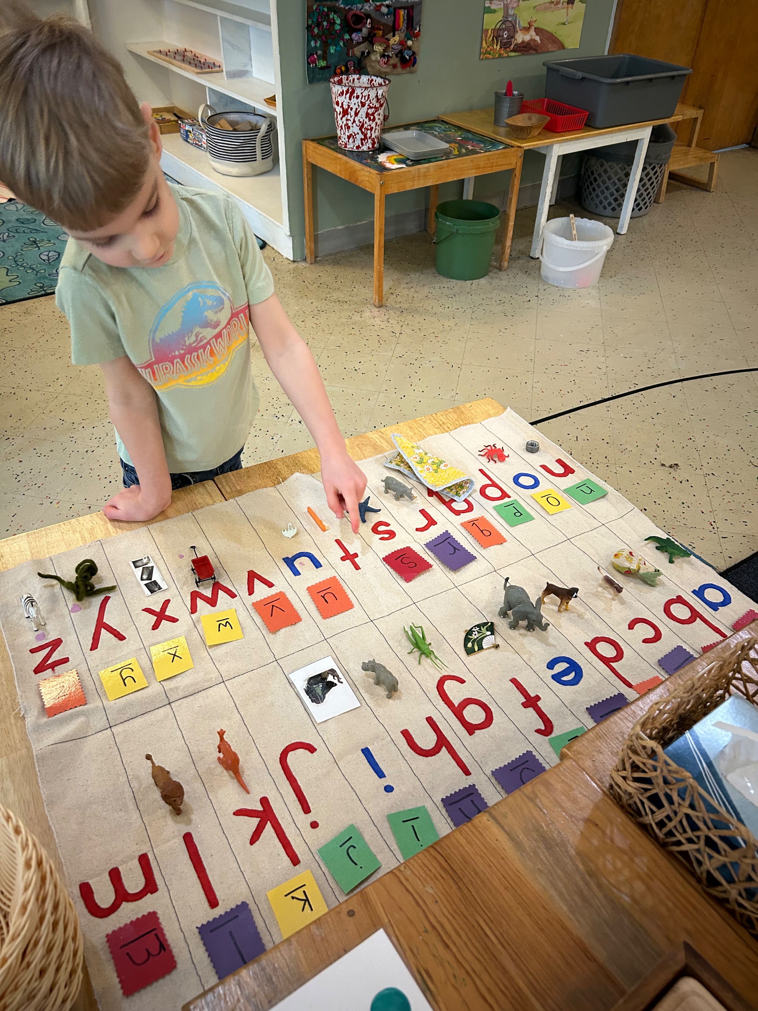 A preschool-aged boy matching small objects to the corresponding letter with which each object starts. Letters are arranged on a large mat on a table.