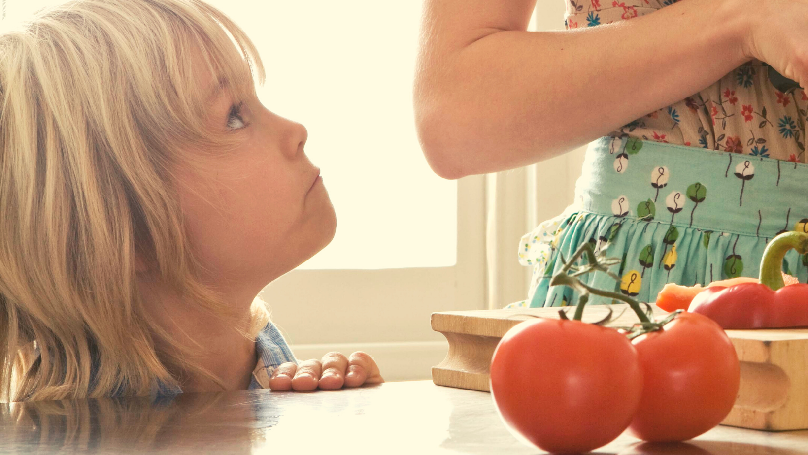 A preschool-aged child looking up at a parent who prepares food. There are tomatoes in the foreground.