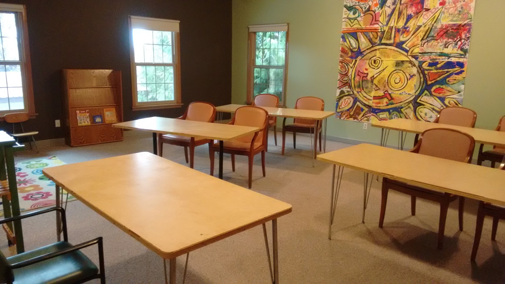 The buildings "Community Room", set up with small tables and chairs, with a large painting of the sun on a wall, and two windows overlooking trees.