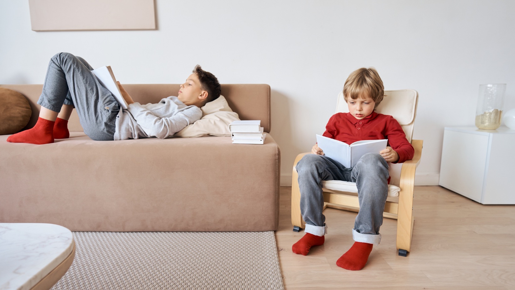 Two elementary-aged boys reading in a family room. On lies on a couch, the other is in a chair.