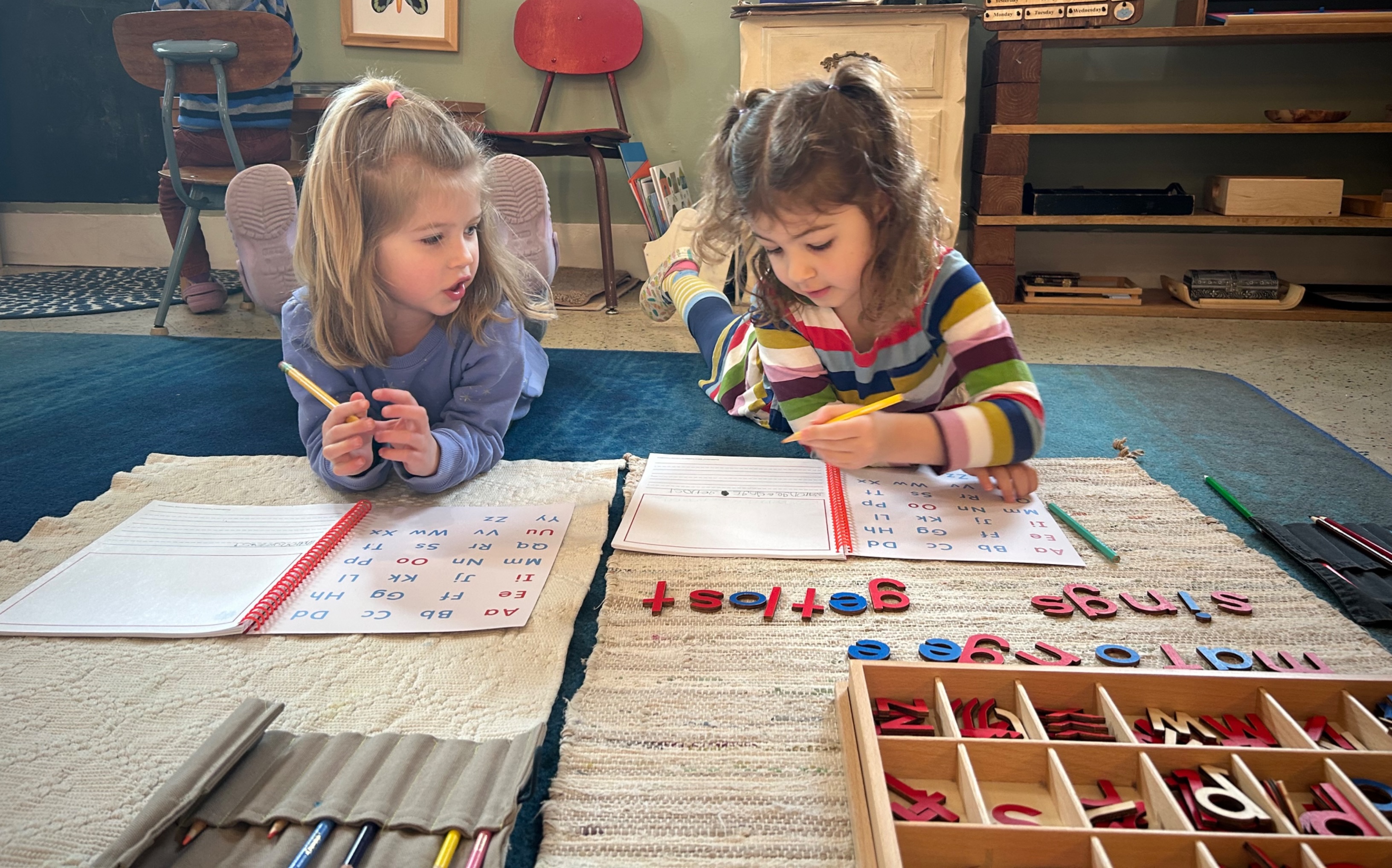 Two preschool-aged girls on the floor using the moveable alphabet and writing in their journals