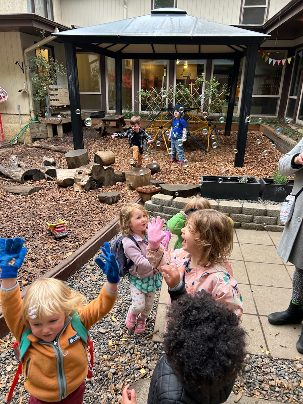 Preschool-aged children joyfully playing with bubbles on the courtyard playground.