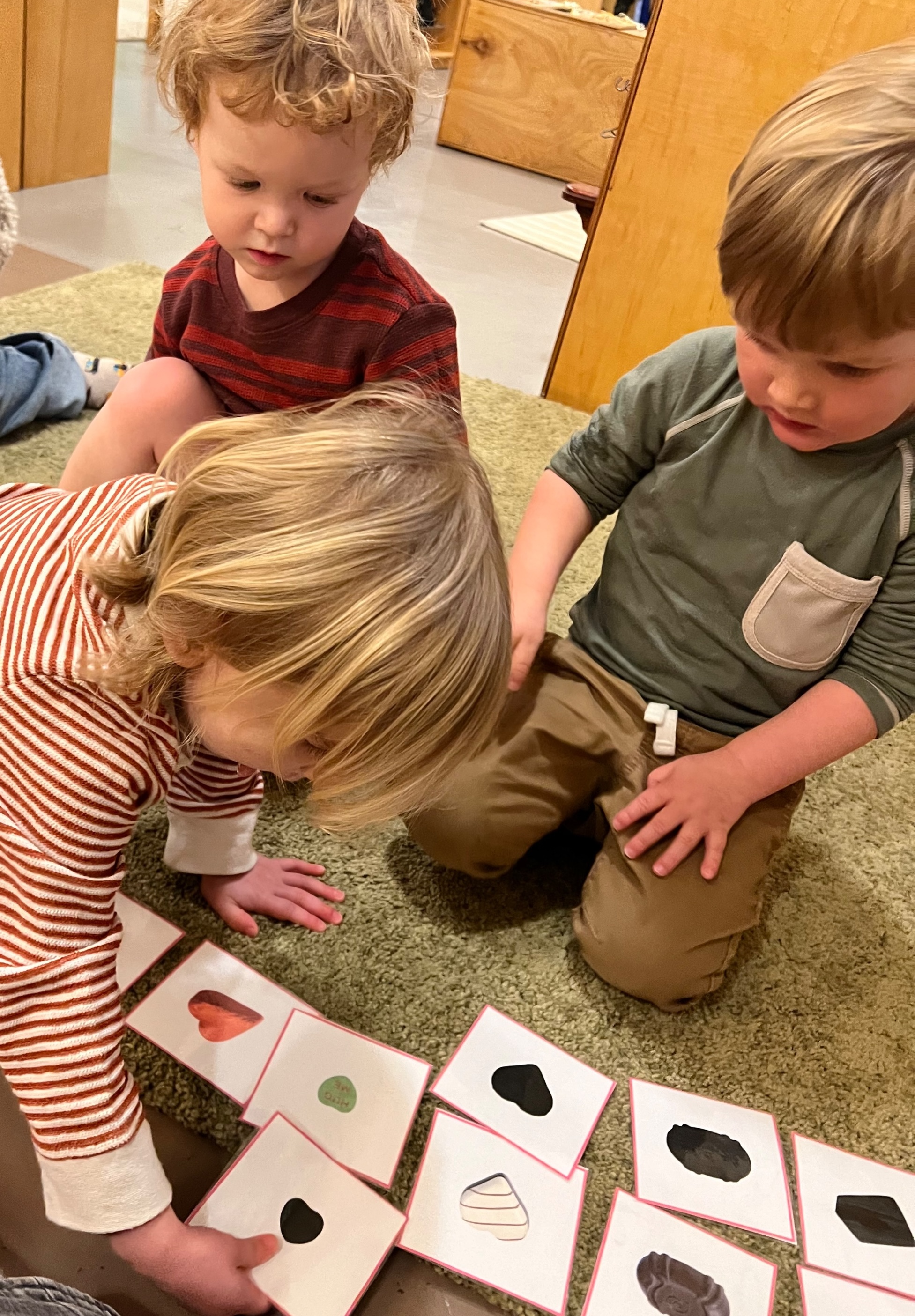 A toddler boy matches cards with silhouettes to their corresponding image on another card. Two other toddler boys observe.