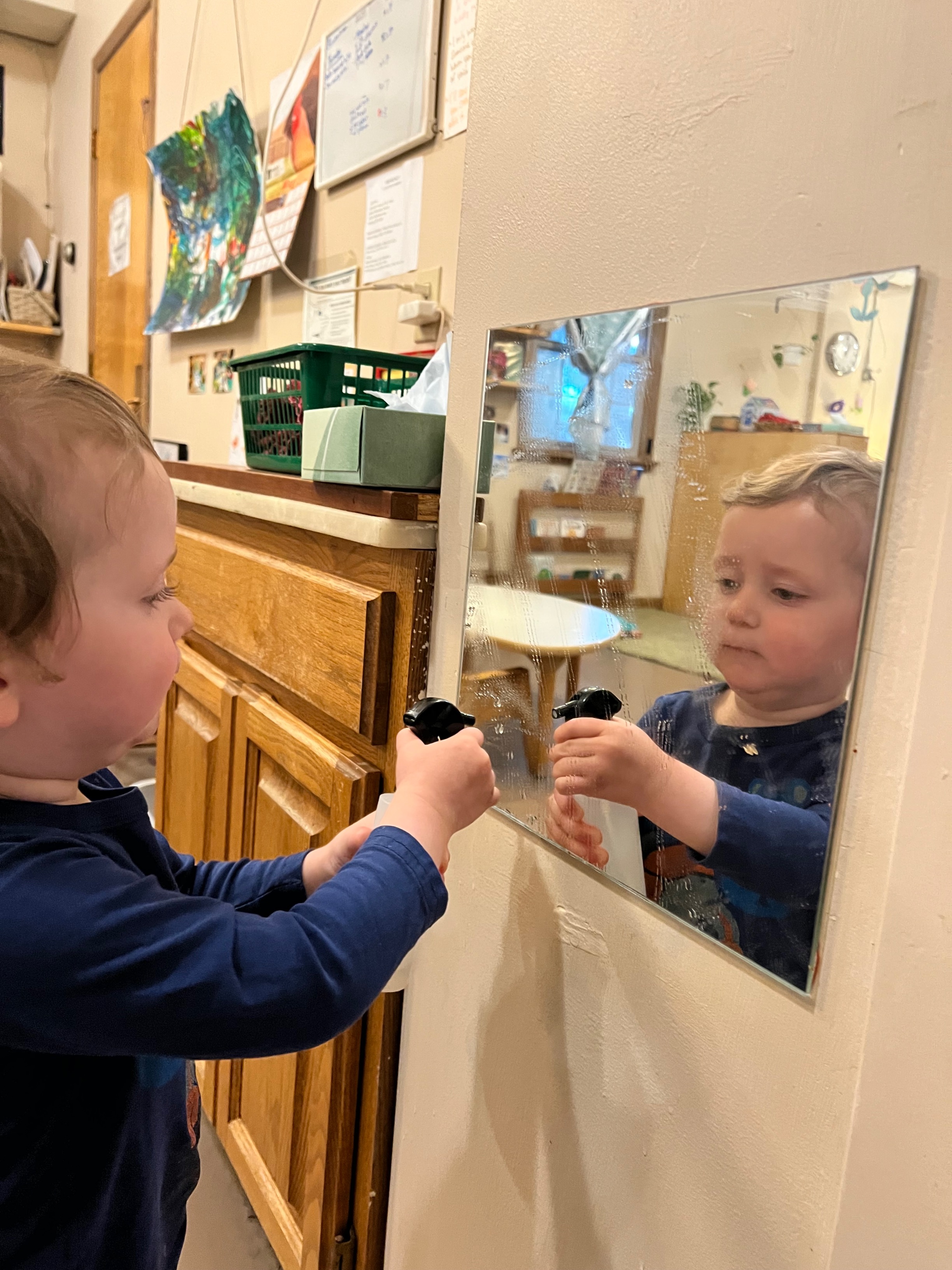 A toddler boy sprays water on a mirror to help clean it.